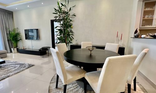 Modern dining room with a round black table, white chairs, and a tall green plant in the corner, adjacent to a living area with a sofa.