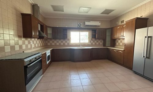 A spacious kitchen with tiled flooring and walls, featuring wooden cabinets, stainless steel appliances, and a central window.