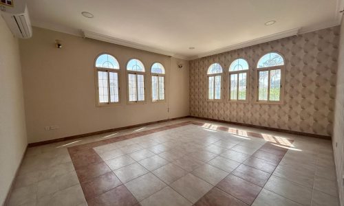 Empty living room with beige walls, arched windows, and tiled floor, sunlight streaming in, creating patterns on the floor.