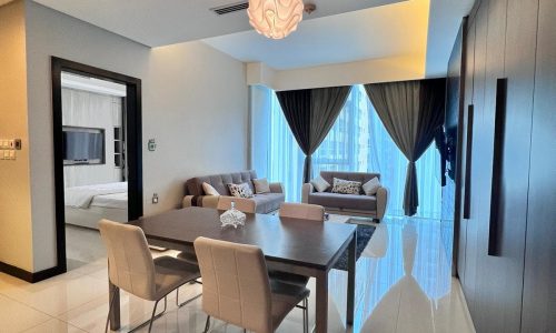 Modern dining room in a luxury apartment with a wooden table, gray chairs, and a stylish pendant light, leading to a living area and visible bedroom.