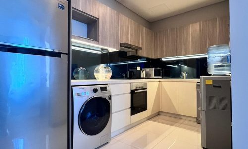 Modern kitchen interior in a luxury apartment with stainless steel appliances, including a refrigerator, oven, and washing machine, under wooden cabinets and white countertops.