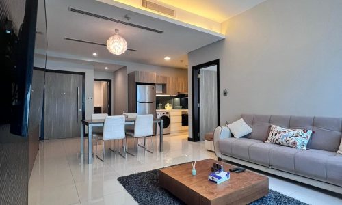 Modern luxury apartment living room with a beige sofa, a wooden coffee table, a black rug, and an open-plan kitchen in the background, illuminated by warm ceiling lights.