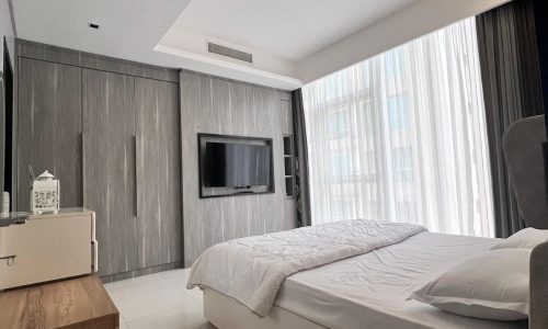 Modern hotel room with a large bed, white linens, and wooden paneling, located in the Juffair area, featuring a mounted TV and sheer curtains filtering natural light.