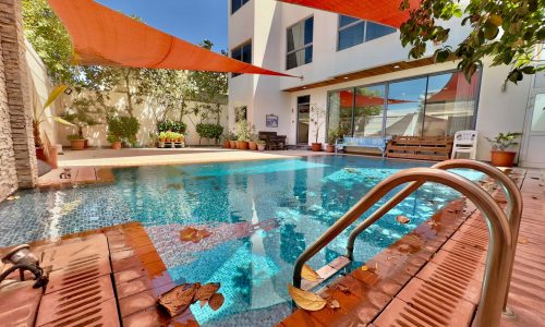 Modern backyard pool with orange sun shades, surrounded by a brick patio, trees, and a white two-story house.