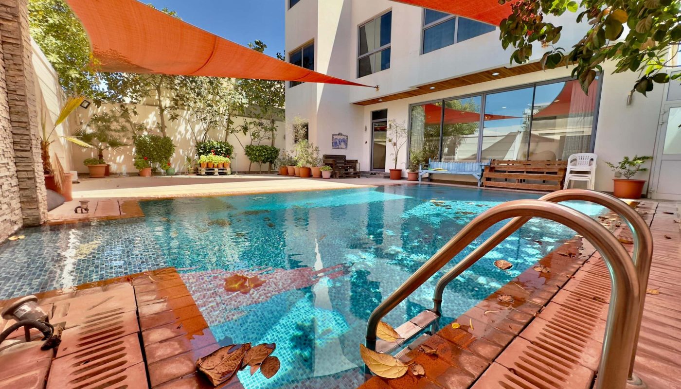 Modern backyard pool with orange sun shades, surrounded by a brick patio, trees, and a white two-story house.