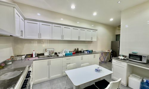 A modern kitchen with white cabinets, gas stove, and a center island, featuring a table with chairs and various household items scattered throughout.