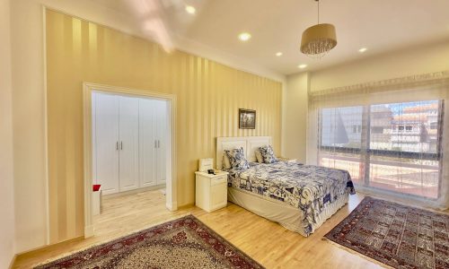 Brightly lit bedroom featuring a double bed with a blue floral comforter, two side tables, a persian rug, and a large window with sheer curtains.