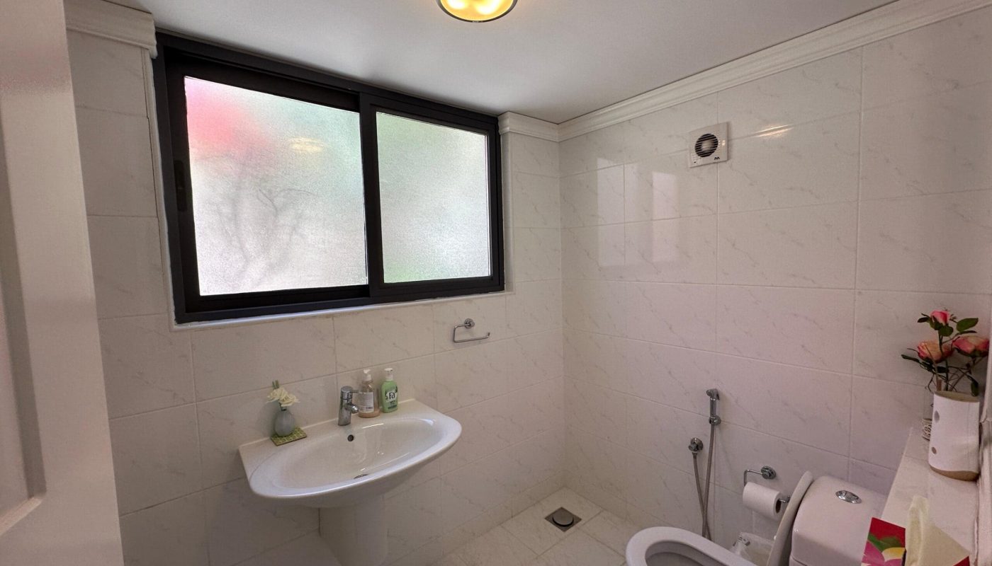 A clean bathroom with white tiled walls, featuring a toilet, a sink, and a frosted window with a small open panel.