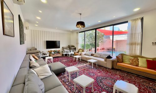 Modern living room with long gray sofa, central coffee tables, persian rug, and large glass window overlooking a patio.