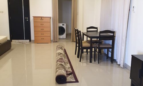 Interior of a modern flat with white walls featuring a rolled-up rug, dining table with chairs, a chest of drawers, curtains, and a washing machine near the entrance.