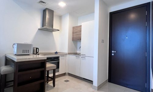 A modern kitchen in an apartment with white cabinetry, stainless steel appliances, and a dark entrance door located in the Juffair area.