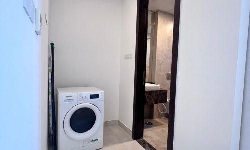 A narrow hallway in a studio for rent in Juffair with a white washing machine on the left and an open door leading to a bathroom on the right.