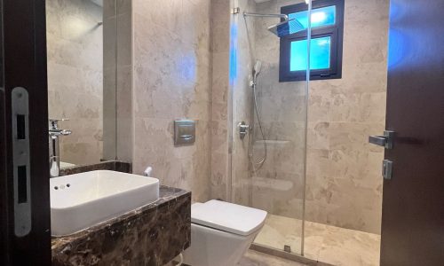 A modern bathroom in a studio for rent in the Juffair area of Bahrain, with beige marble walls, featuring a bathtub, toilet, shower area, and a small blue-tinted window.