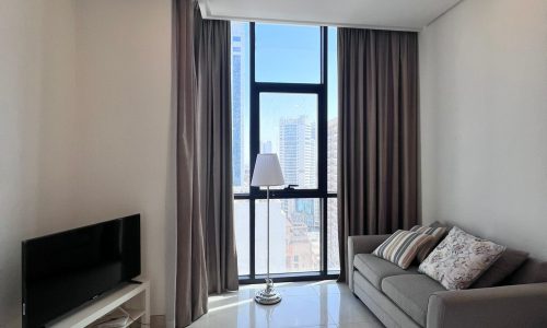 Modern living room in a studio for rent with a gray sofa, TV on a white stand, floor lamp, and large window with city view in the Juffair area, featuring minimalistic decor.