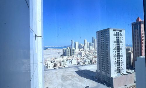 View of the Juffair area cityscape with buildings under construction and a parking lot, seen through a blue-tinted window.