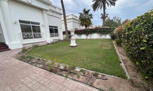 A lush garden with trimmed grass and flowering bushes next to a luxury villa with ornate windows and a sundial sculpture, under a cloudy sky.