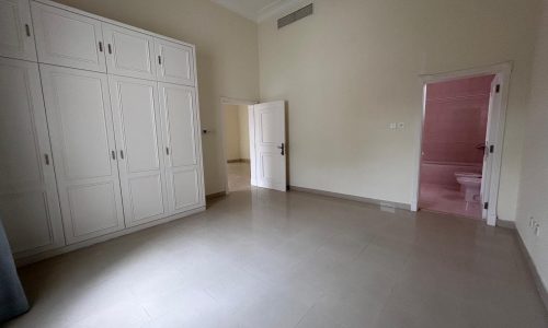 Empty room in a Saar Villa with white walls, large white wardrobe, tiled floor, and two doors, one leading to a pink bathroom.