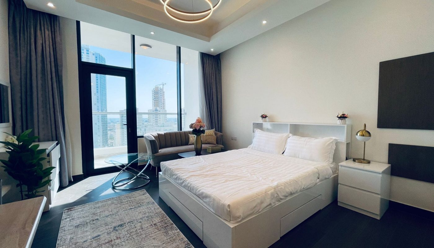 Modern bedroom in a luxury studio with a double bed, white bedding, and side tables, featuring a balcony view of high-rise buildings in Juffair.