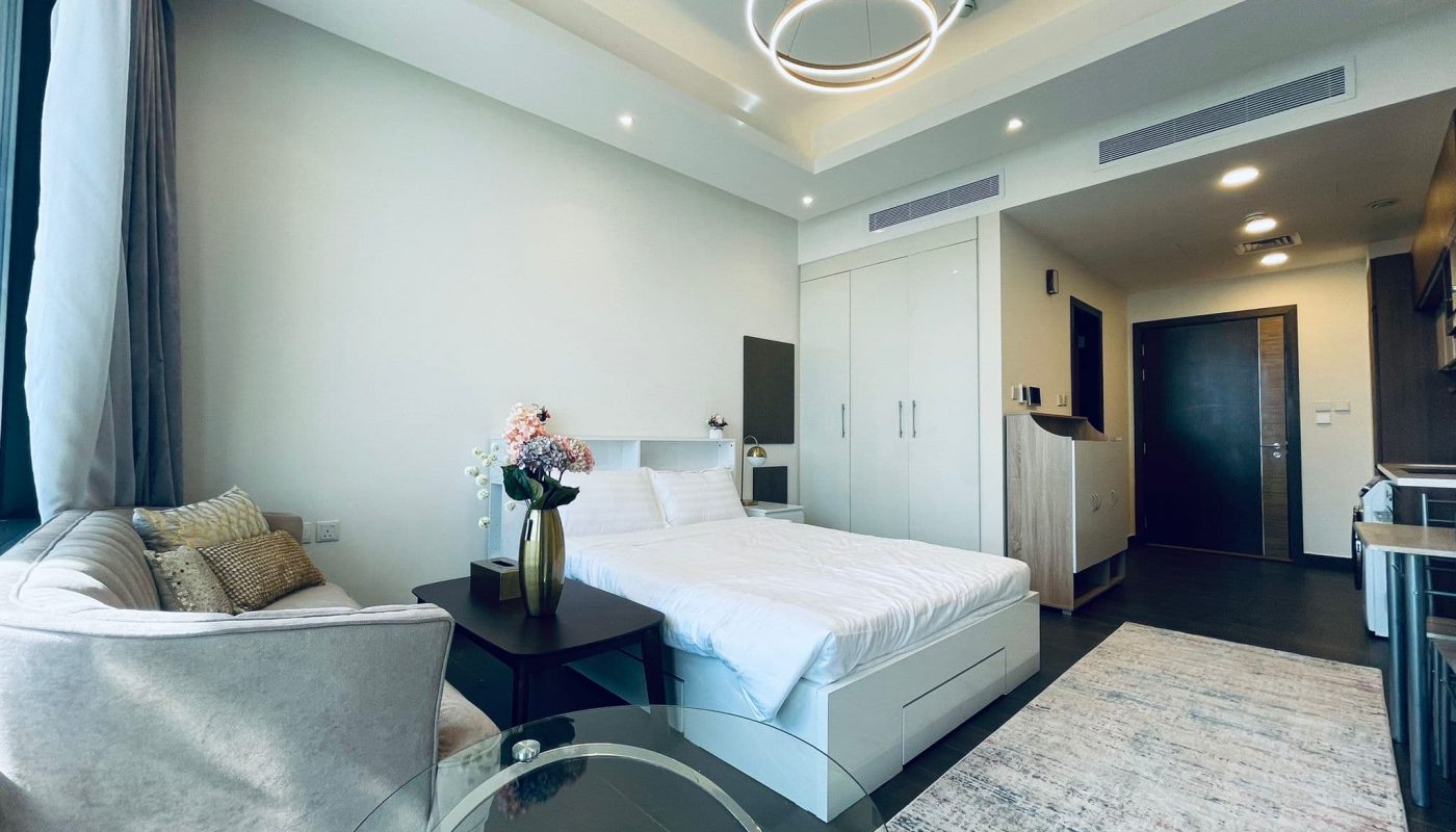 Luxury studio apartment interior in Juffair with a bed, sofa, glass coffee table, rug, and elegant lighting fixture. Light decor with a balcony door.