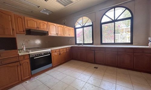 An empty kitchen with wooden cabinets, a built-in oven, and a large arched window overlooking lush greenery.