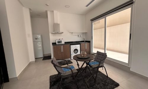 A small, modern kitchen with appliances and a dining area featuring a table, two chairs, and an auto draft rug, beside a large window and a sliding door.