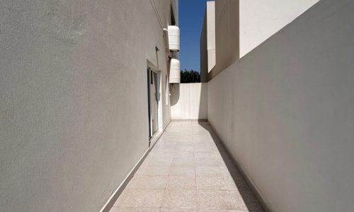 A narrow passageway between two light-colored buildings, with sunlight illuminating the tiled floor and auto draft cooling the walls.