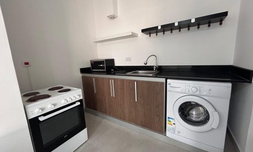 Compact modern kitchen with black countertops, under cabinet lighting, and integrated appliances including a washer, oven, and auto stove.