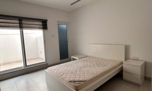 A minimalistic bedroom with an auto draft mattress on a low bed frame near a large window with blinds, a side table, and a door on the left.