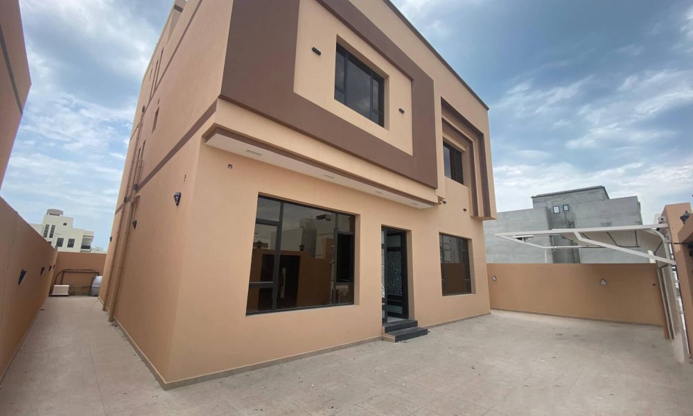 A two-story beige house with dark brown accents, large windows, a small staircase leading to the entrance, and an open concrete courtyard under a cloudy sky emulates the sophistication of an elegant villa in the Danat Hidd scheme.