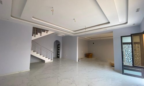 Spacious empty room with marble floors, a staircase, and a high ceiling with recessed lighting in an elegant villa in Danat Hidd. One wall features a sliding glass door with a decorative pattern.