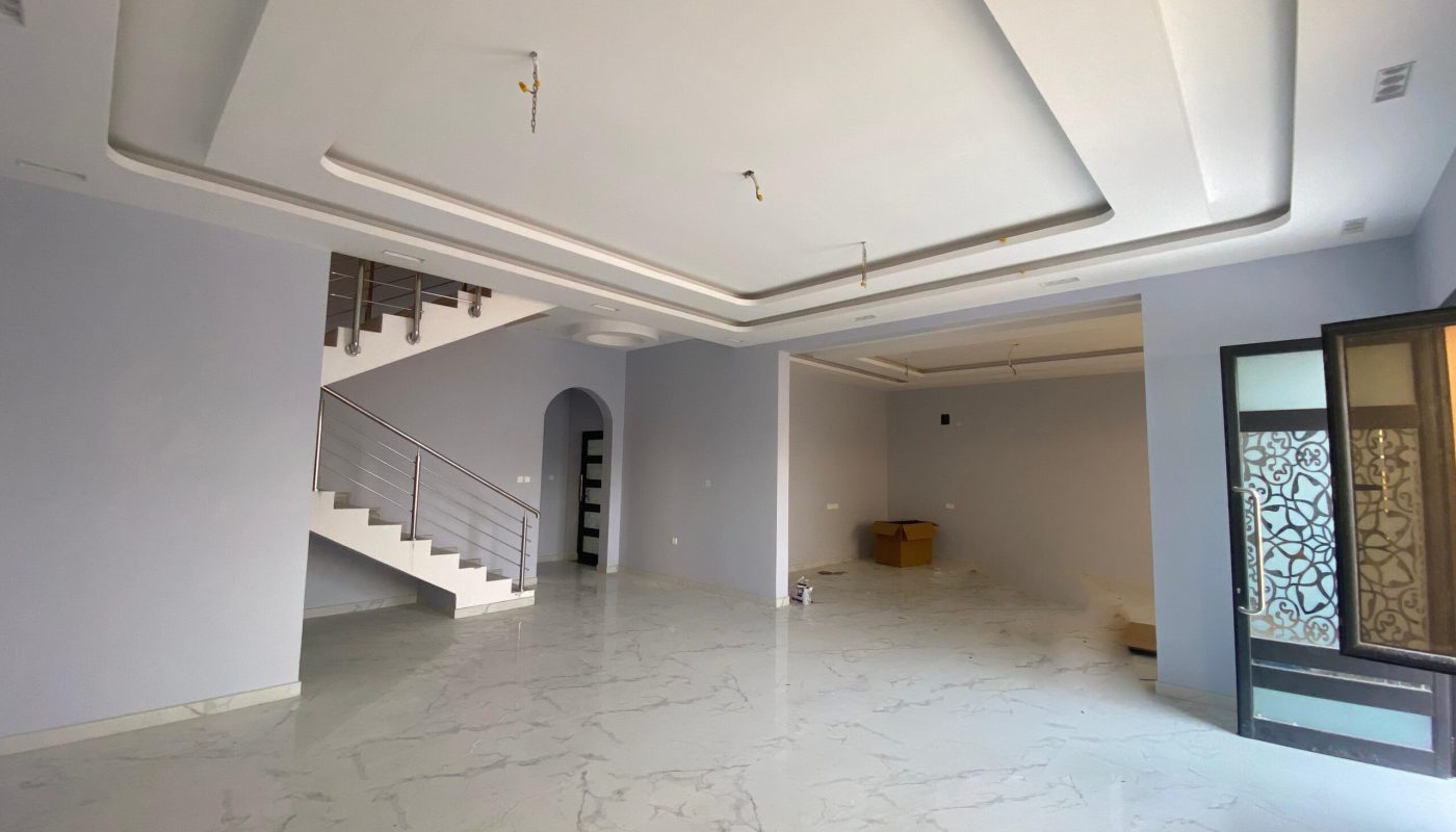 A spacious, unfurnished room with white marble floors, an ornate ceiling, a staircase, and several doorways and windows in an elegant villa in Hidd.
