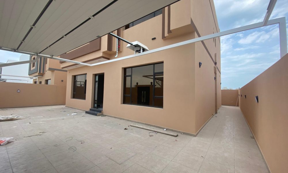 A beige two-story house with large windows and an open garage area stands proudly in the Danat Hidd scheme. The exterior is clean against the clear sky, exuding the charm of an elegant villa. Some construction materials and debris are visible on the ground.