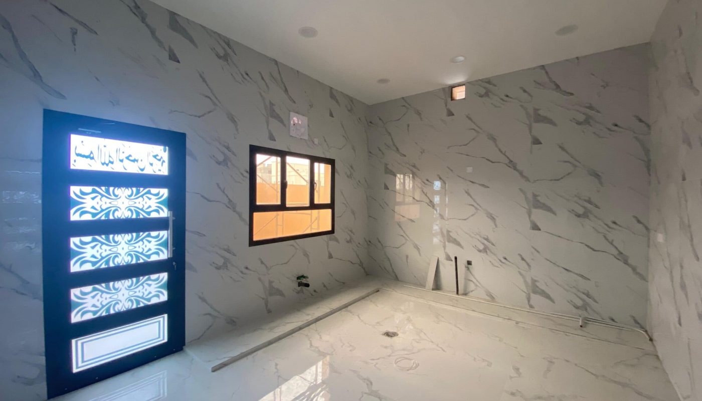 A room with marble-patterned walls and floor, a decorative door, a window, and light fixtures on the ceiling. Within this elegant villa in the Danat Hidd scheme, construction materials are scattered on the floor.