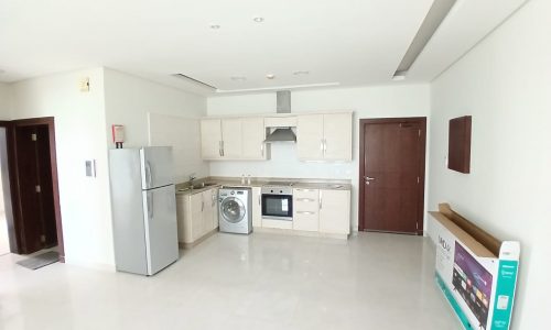 A modern kitchen in a Flat for Rent with white walls featuring a refrigerator, oven, washing machine, and wooden cabinets.