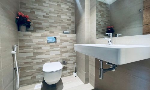 A modern bathroom with beige tiled walls, featuring a white toilet, wall-mounted sink, and a small shelf with flowers.