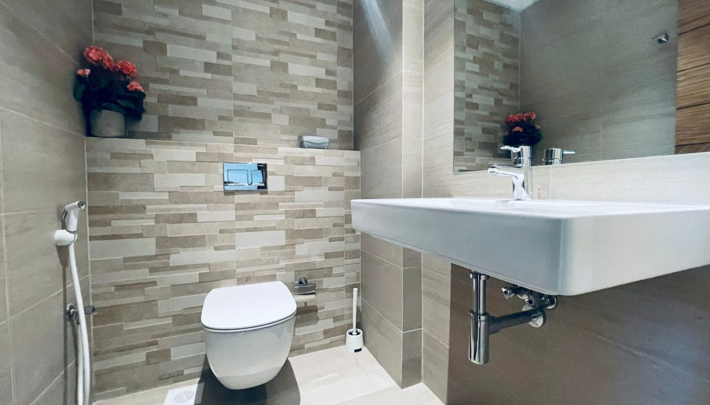 A modern bathroom with beige tiled walls, featuring a white toilet, wall-mounted sink, and a small shelf with flowers.
