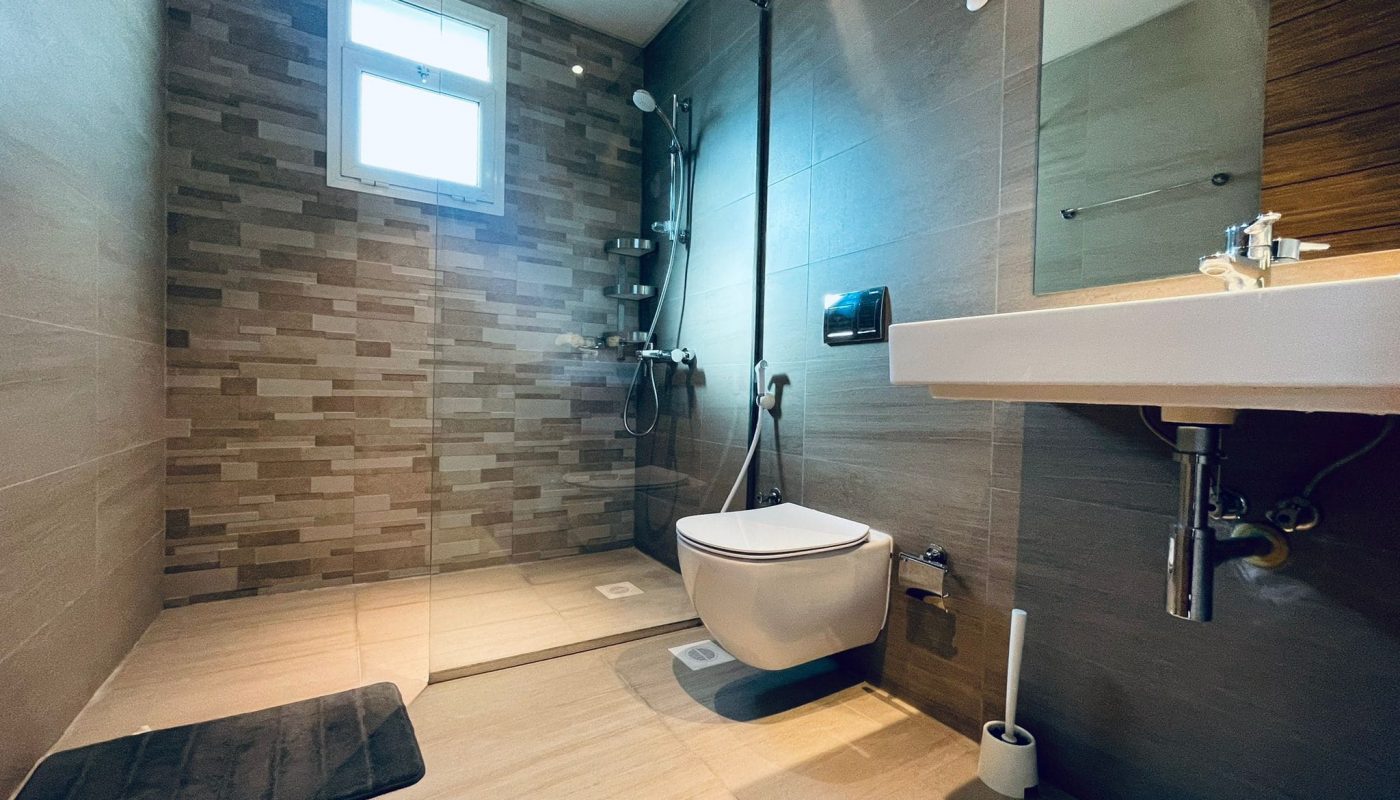 Modern bathroom with a floating toilet, glass shower area, tiled walls, and a wall-mounted sink under a small window.