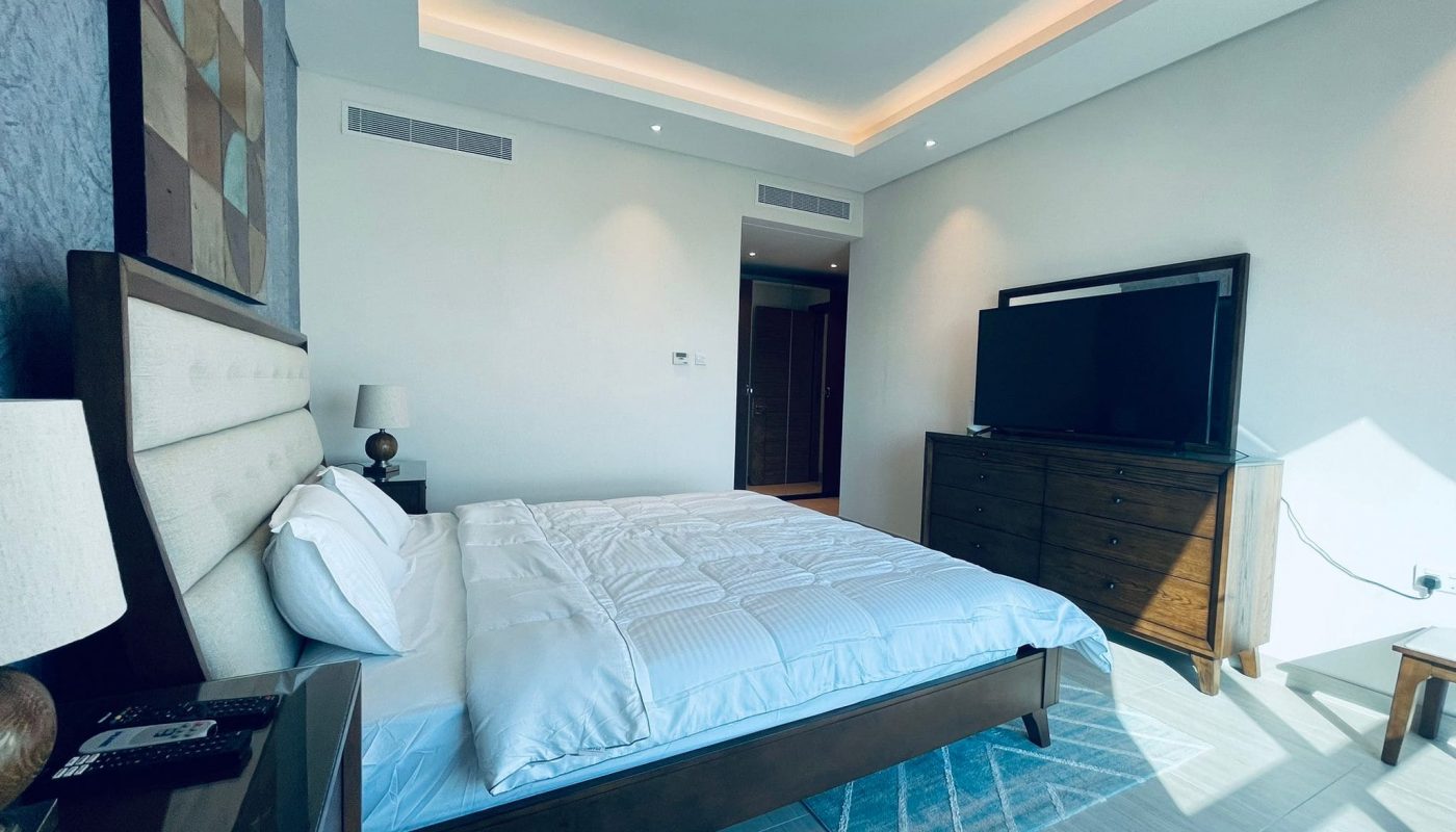 Modern bedroom with a large bed, white bedding, wooden side tables, lamps, a dresser, and a flat-screen tv, under soft ambient lighting.