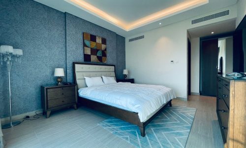 A modern bedroom with a king-sized bed, gray textured walls, and ambient ceiling lighting.