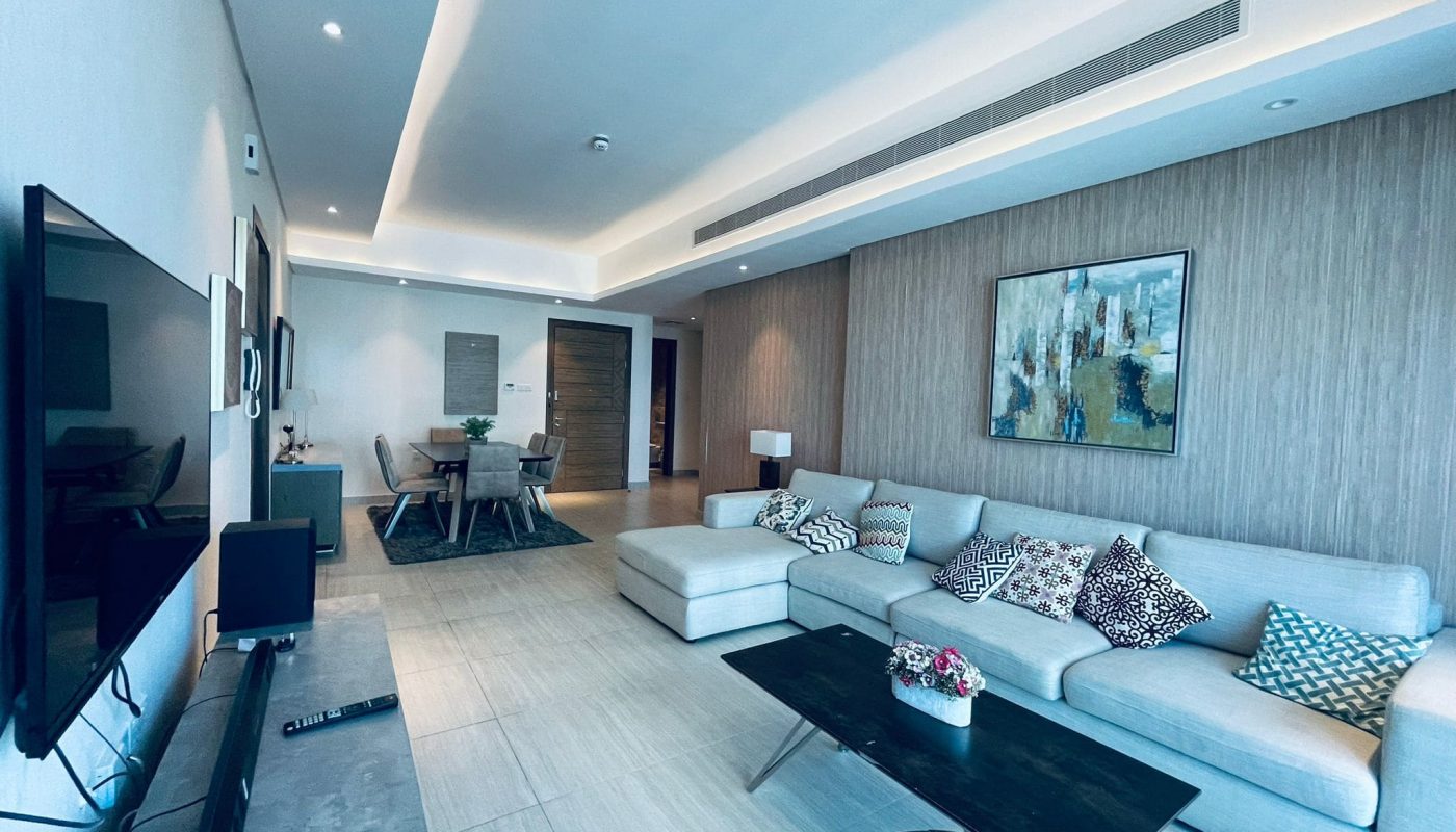 Modern living room with a light gray sectional sofa, patterned cushions, a glass coffee table, and a large wall-mounted TV featuring neutral colors and auto draft.