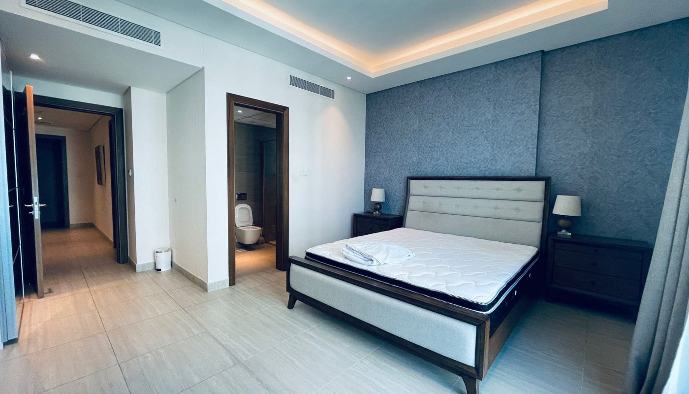 Modern bedroom with a large bed, blue accent wall, ambient lighting, and an open door leading to an auto draft bathroom.