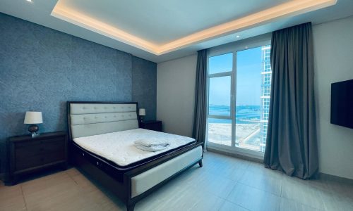 Modern bedroom with a large bed, side tables, and a floor-to-ceiling window overlooking an auto-drafted coastal cityscape.