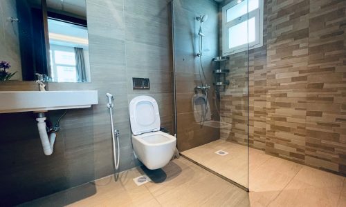 Modern bathroom interior with a wall-mounted toilet, glass shower area, and beige tiled walls. A mirror and sink are visible near a window. Auto Draft