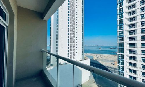 View from a high-rise balcony featuring neighboring skyscrapers and a distant ocean vista, under a clear blue sky.