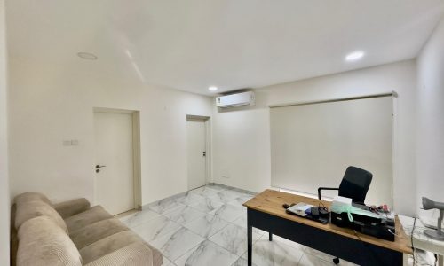 Modern office with a beige sofa, a wooden desk with items, an air conditioner, white walls, and tiled floors. two doors are visible on the left side.