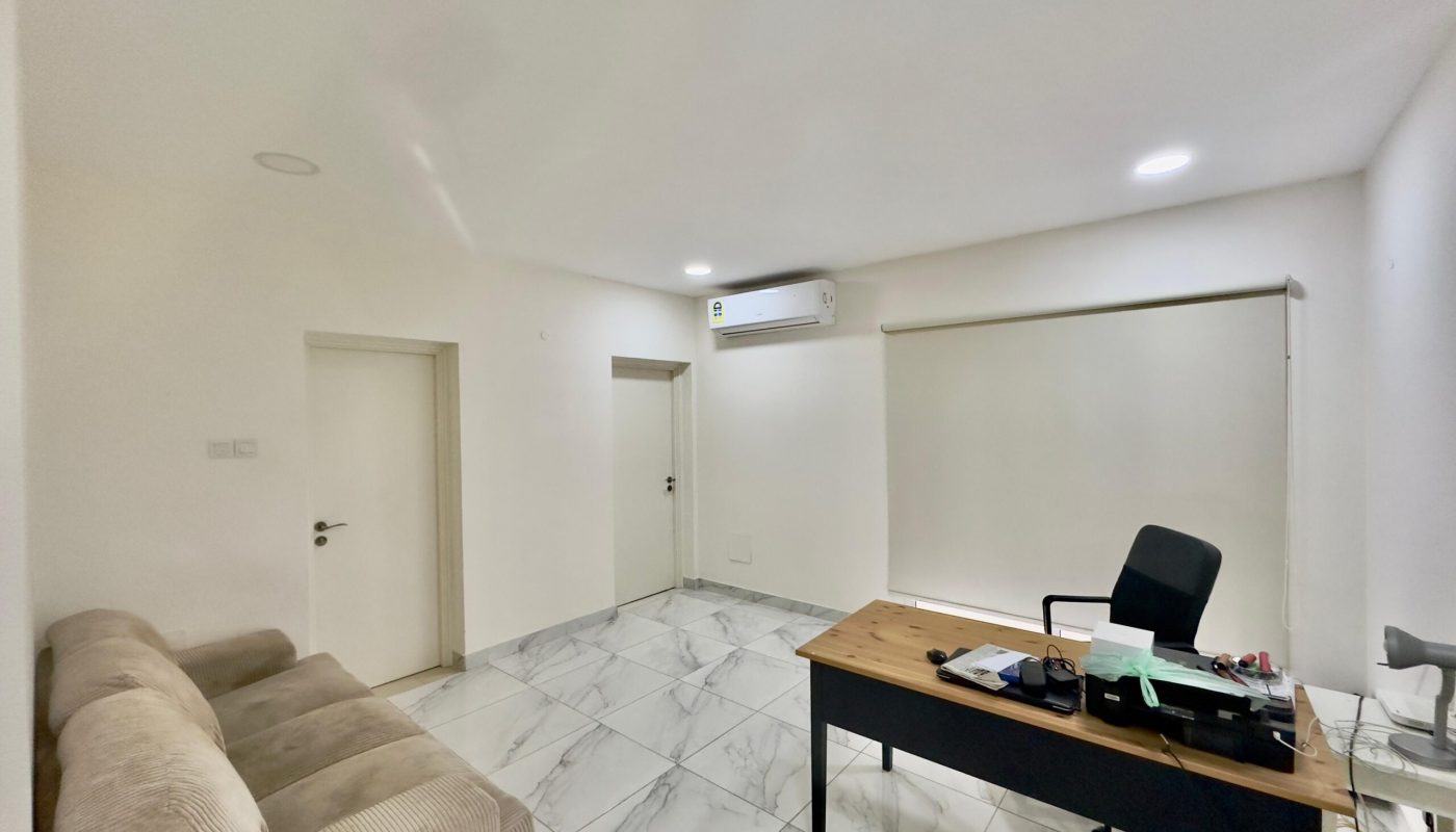 Modern office with a beige sofa, a wooden desk with items, an air conditioner, white walls, and tiled floors. two doors are visible on the left side.