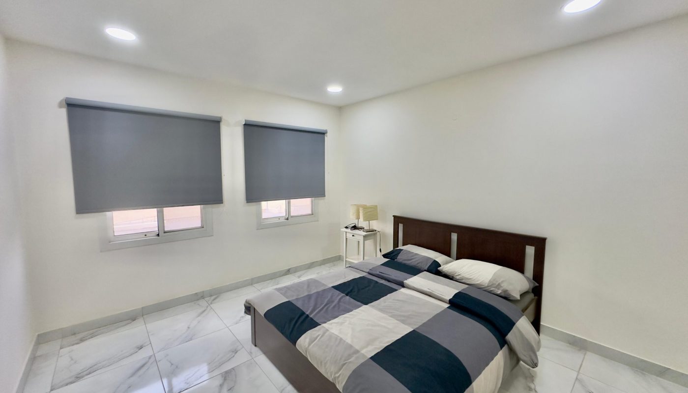 A modern bedroom featuring a double bed with striped bedding, two gray roller blinds on the windows, and glossy tile flooring.