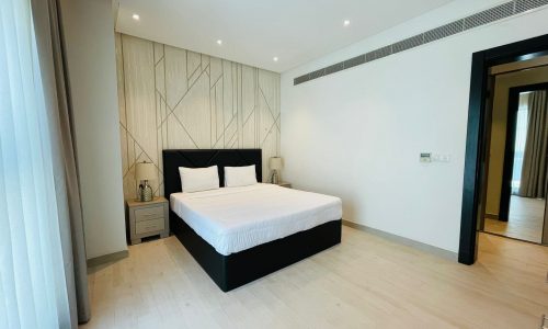 Modern bedroom in a stylish apartment with a large double bed, white bedding, wooden headboard, and patterned wall panels, featuring minimalistic decor and natural light.