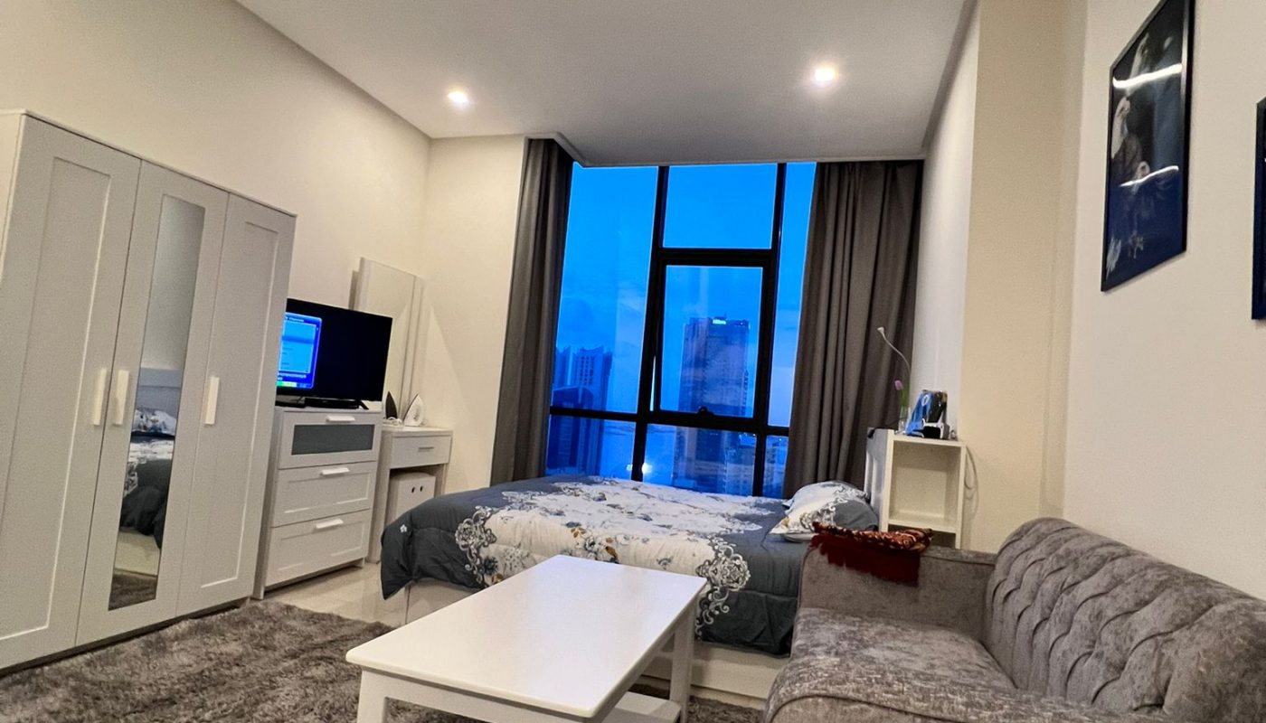 A modern living room with a gray sofa, white coffee table, and a tv. large windows provide a view of the evening sky.