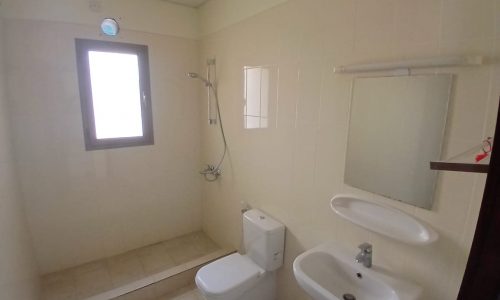 A small, basic bathroom with a toilet, sink, shower, mirror, and window. the walls are tiled and painted a light color.